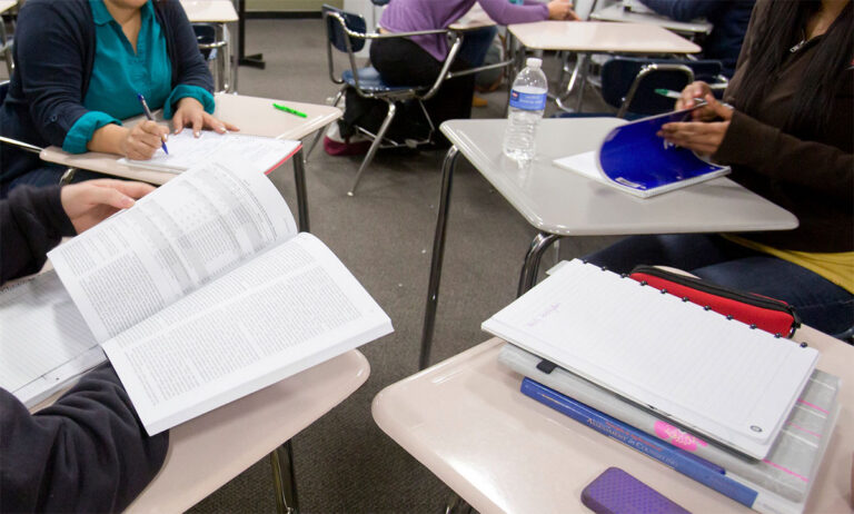 Four classroom desks, and students looking at notebooks and materials