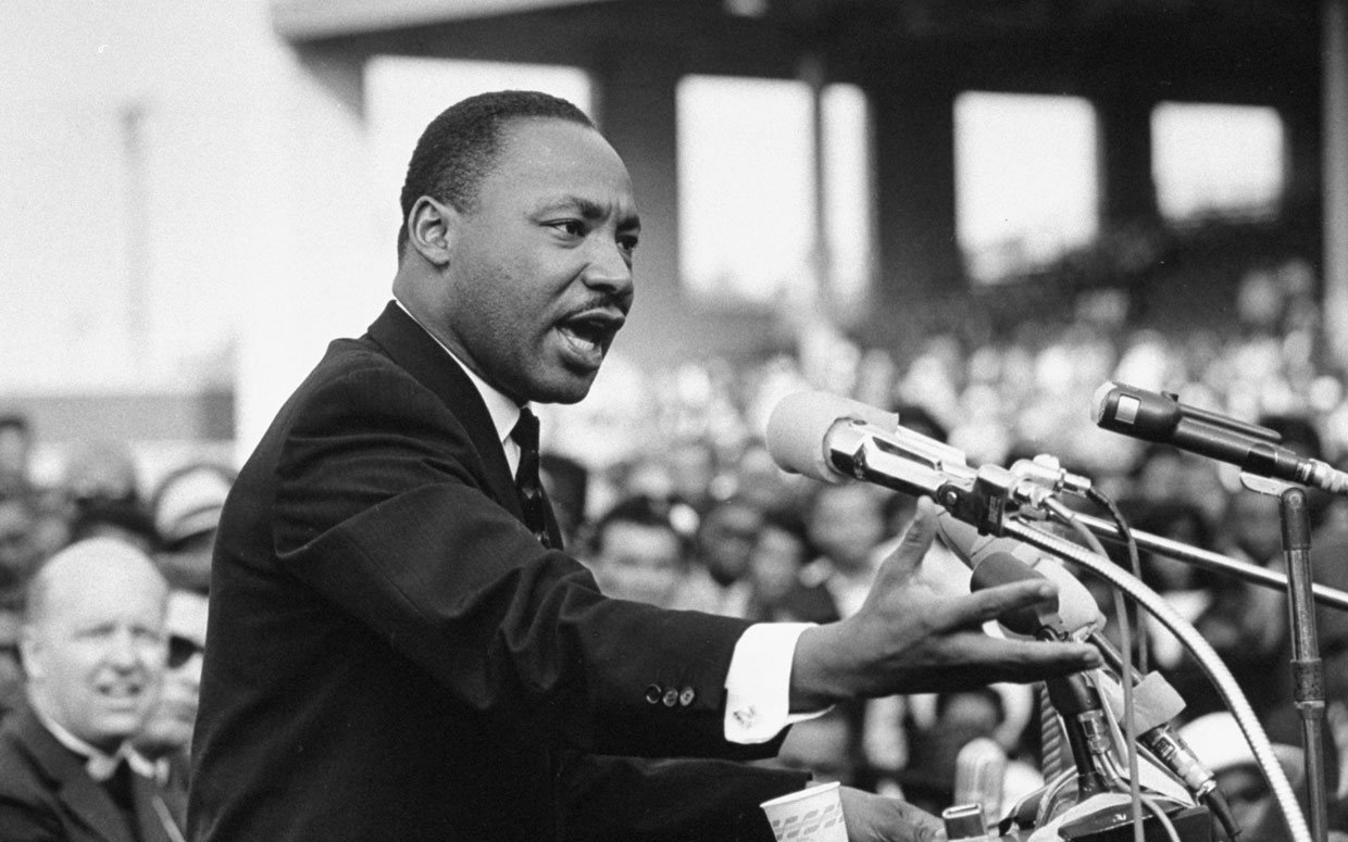 Dr. Martin Luther King, Jr. speaks at a podium outside to a crowd.