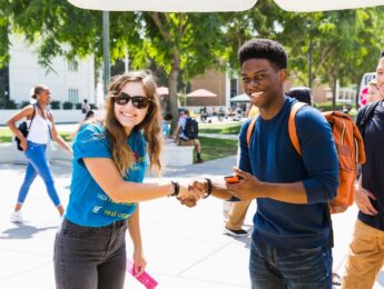 Two students shaking hands outside on campus during an event, one student is wearing a blue t-shirt and the other is wearing a blue sweater with a brown backpack.