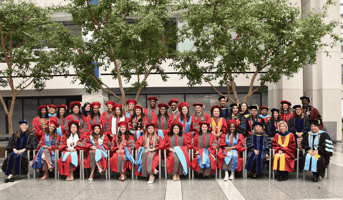 Doctoral graduates and faculty wearing commencement regalia and sitting in an indoor courtyard