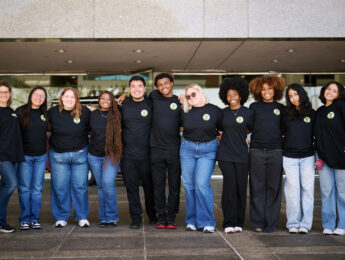 Students pose for a group photo outside of glass doors wearing jeans and black t-shirts.