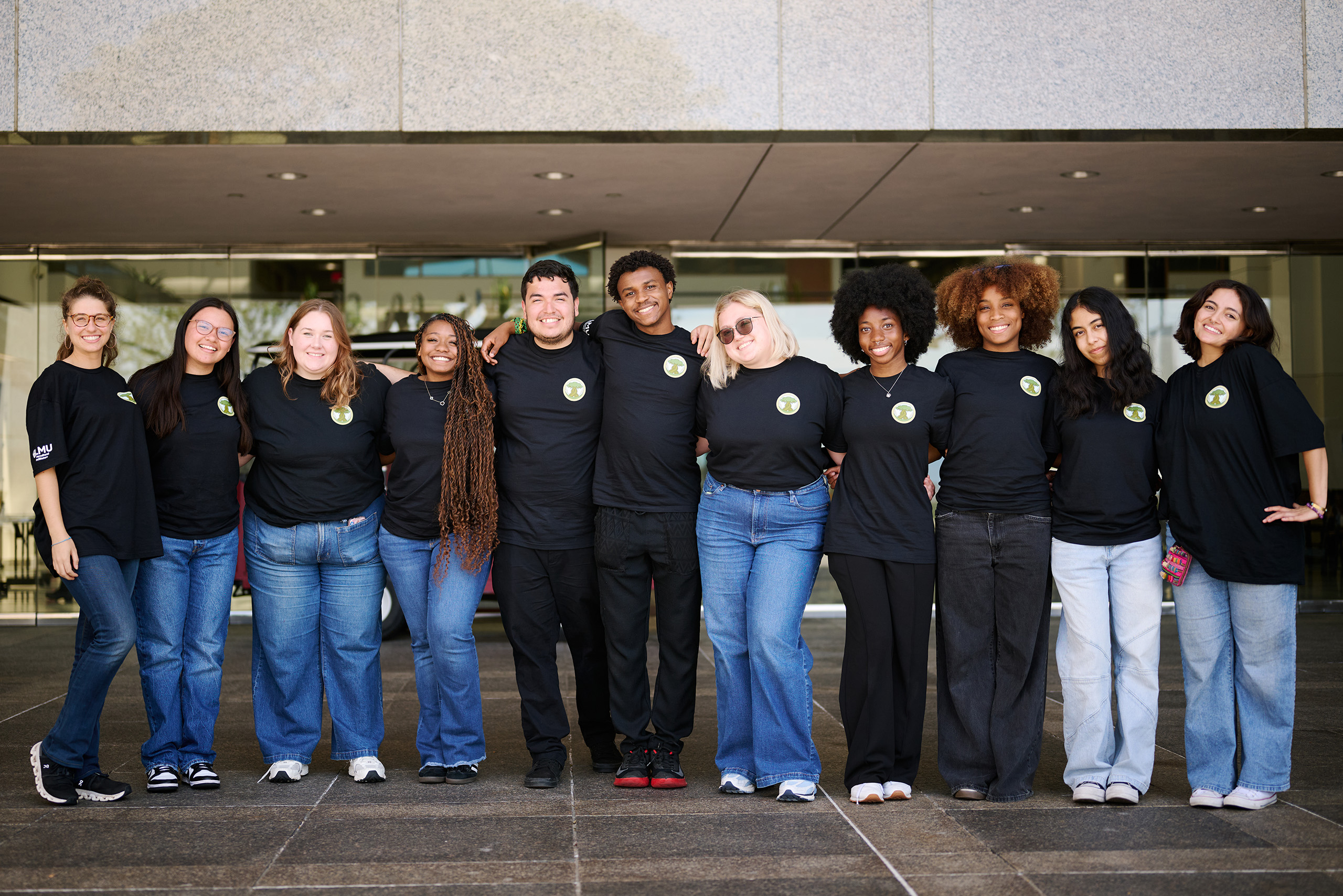 Students pose for a group photo outside of glass doors wearing jeans and black t-shirts.
