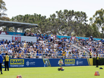 Green grass with people filling stands outside watching a football practice.