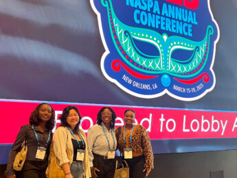 Four students stand inside a convention center with blue and red neon signs in the background.