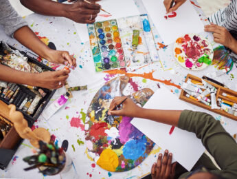 Ariel view of a table covered in art supplies and a few kids arms working on their paintings