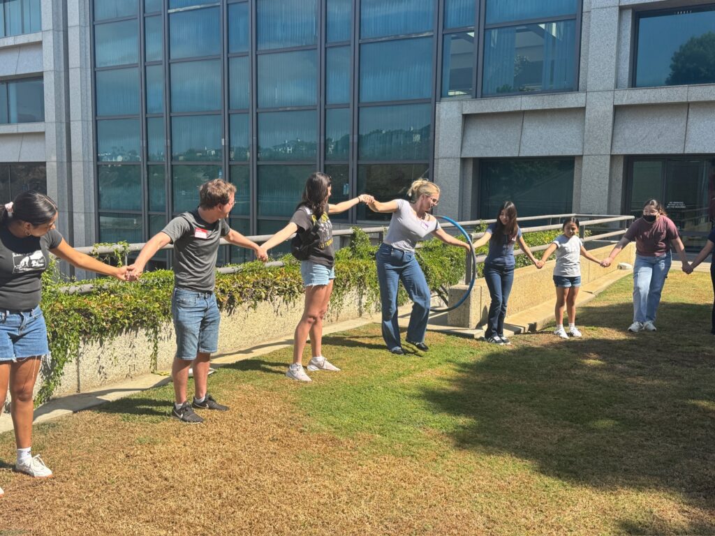 Students stand in a line holding hands as they try to pass a hula hoop around the circle.