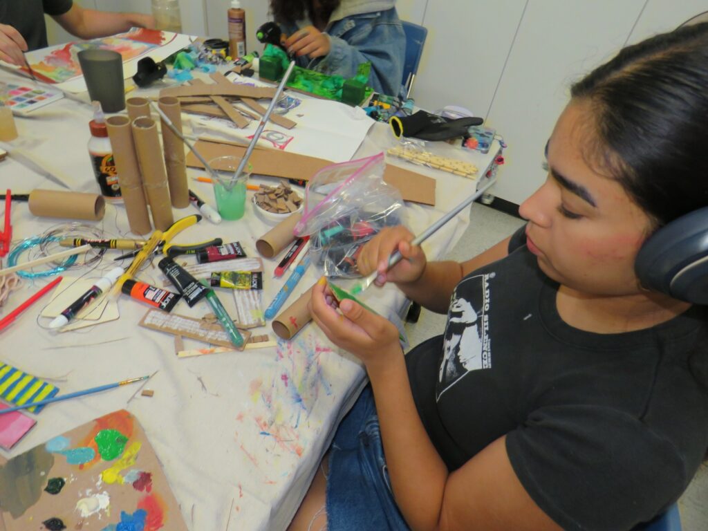 A girl sits at a table with art supplies all around her. She paints.