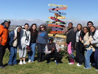 Students and a staff member pose outside with a colorful sign with arrows in South Africa.