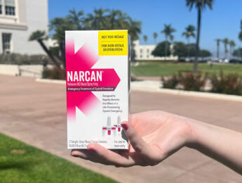 A student's hand holds a pink and white box with Narcan on the package.