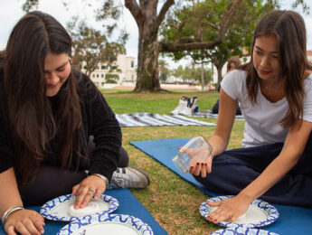 Two students sit on the ground molding clay with their hands outside.