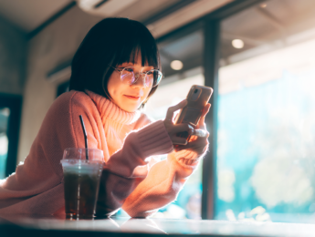 A woman wearing glasses and a pink sweater sits by a café window, smiling as she looks at her smartphone. A cold drink with a straw rests on the table beside her, while warm sunlight streams through the window.
