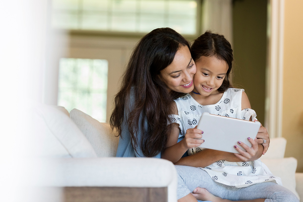 A mid adult mother holds her elementary age daughter in her lap. They look down at a digital tablet as they sit in their living room together.