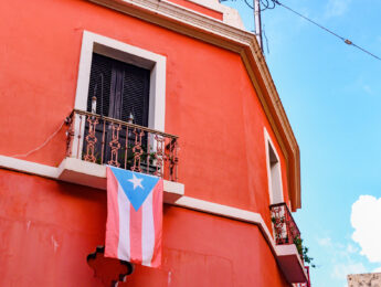This is a color photograph of a Puerto Rican flag hanging from the balcony of a red building in Old San Juan, Puerto Rico.