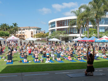 Students stretch on yoga mats outside on Lawton Plaza led by an alum instructor with the Hannon Library and blue skies in the background.