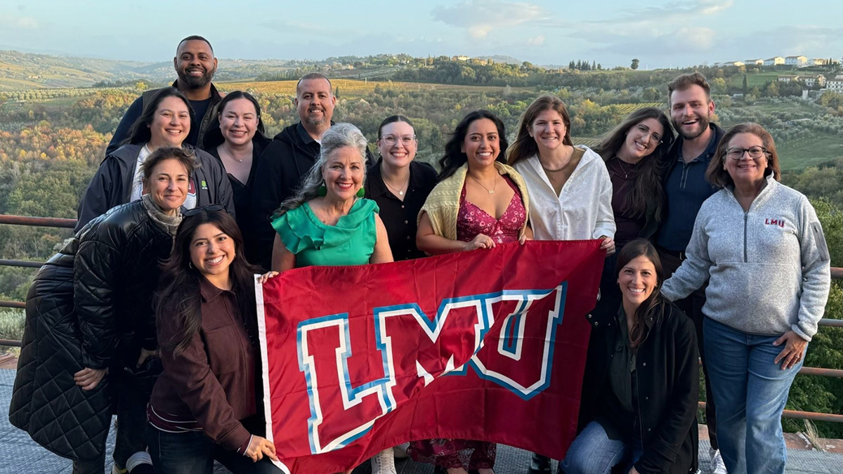 Alumni pose with a red, white, and blue LMU flag with the landscape of Italy in the background