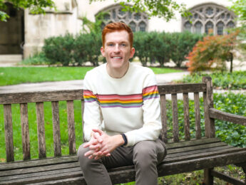 Prof. Hansen sitting on a bench surrounded by greenery wearing a white sweater with an old building in the background.
