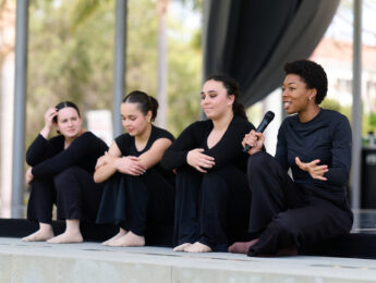 Four dancers sit on stage; one talks into a microphone