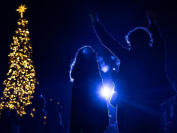 A dark nighttime view of two students in shadow in front of a spotlight during the annual Tree Lighting for Christmas.