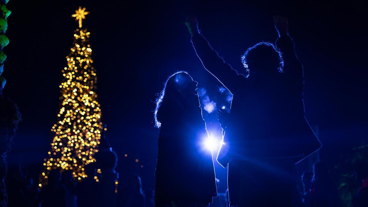 A dark nighttime view of two students in shadow in front of a spotlight during the annual Tree Lighting for Christmas.