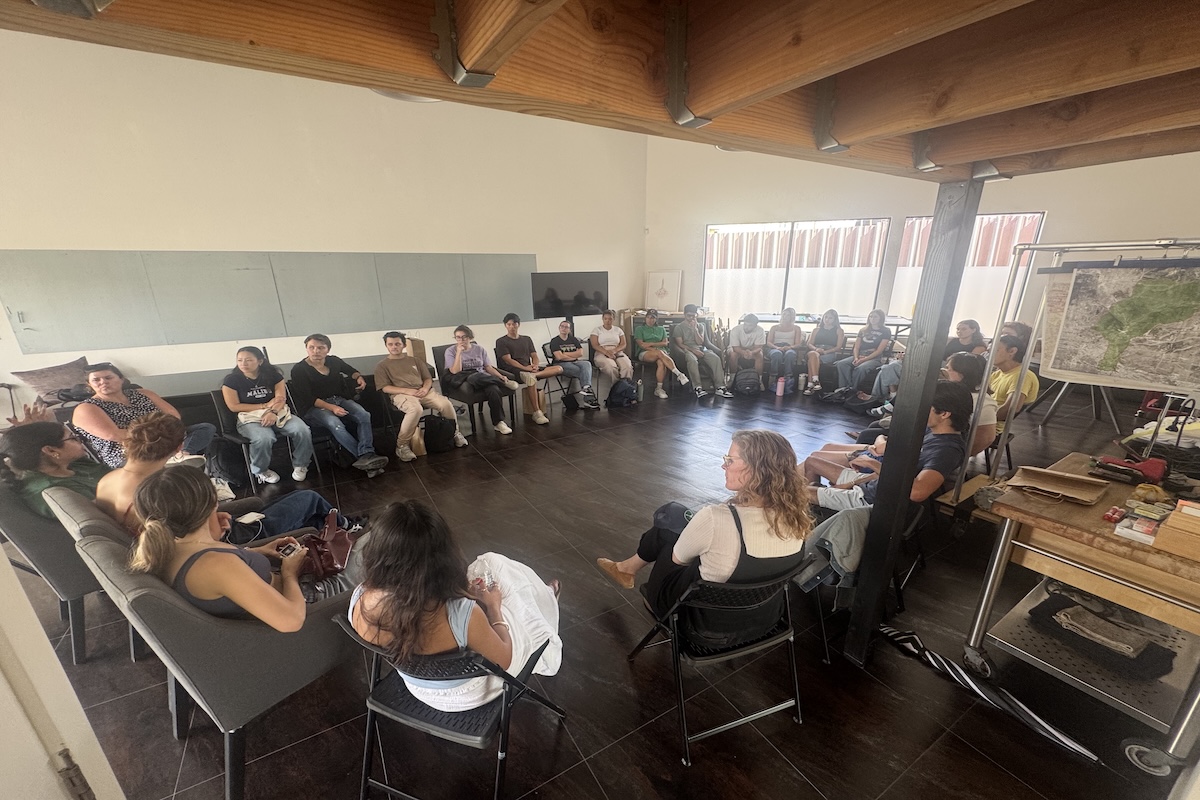 Students sitting in circle engaged in conversation in room with wood paneled ceilings.