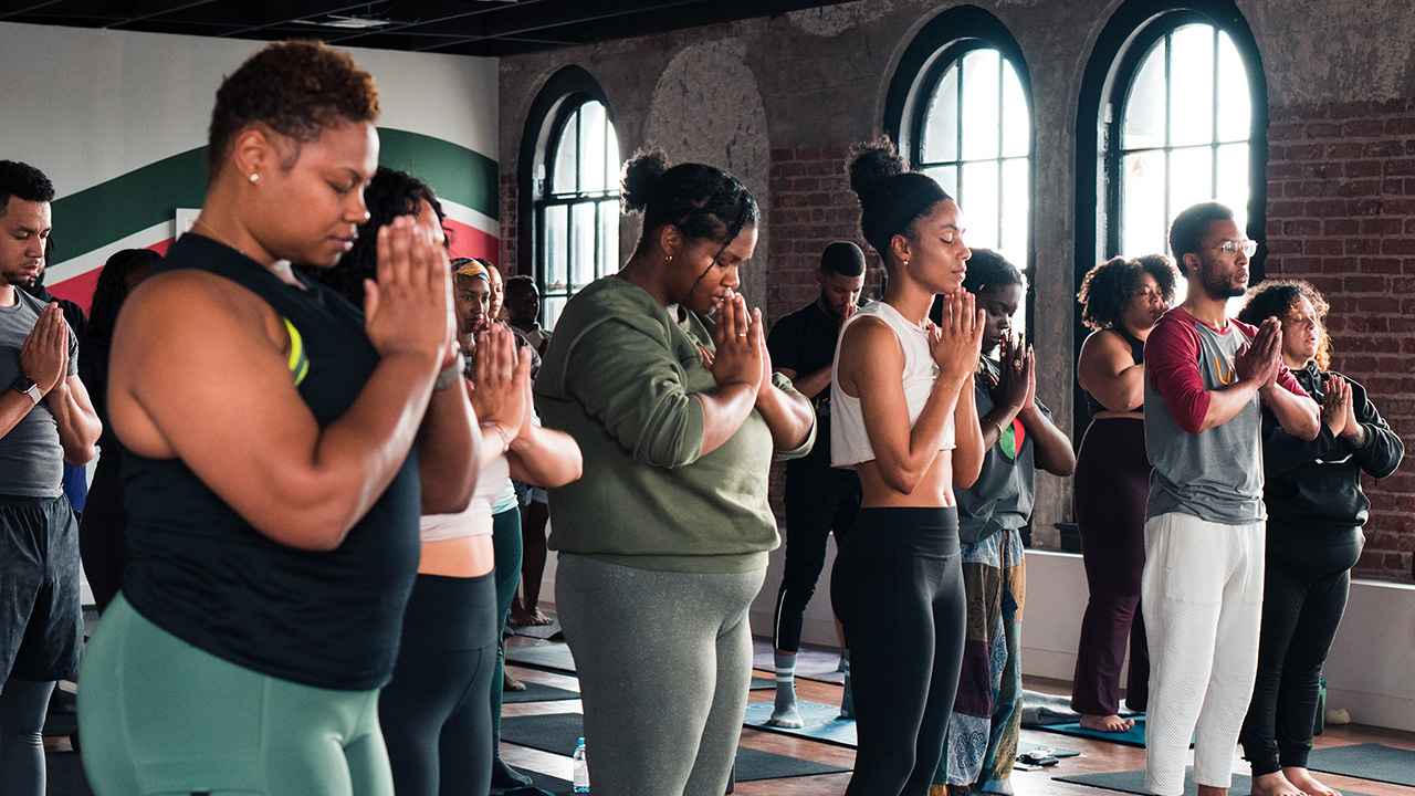 People stand posing during a yoga class with their hands in a prayer position and eyes closed.