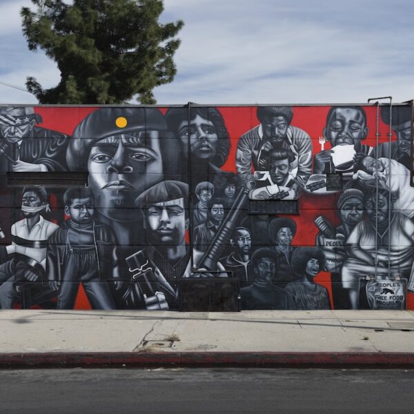 Mural in black, white and red depicting faces and figures of people on the side of a building. Image credit: Noni Olabisi, "To Protect and Serve," 1995, Acrylic, 12 x 40 ft., 11th Ave. at Jefferson Blvd. Photo credit: Social and Public Art Resource Center (SPARC), The Great Walls Unlimited: Neighborhood Pride Mural Program, 1988-2002, Courtesy of Laband Art Gallery, Copyright held by the Estate of Noni Olabisi