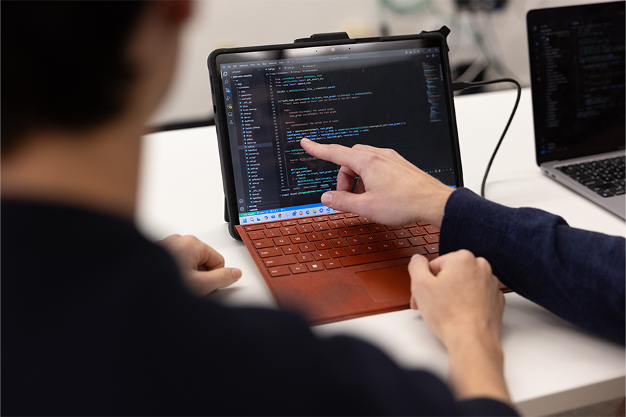 Jared Coleman's hand pointing to a computer screen with code as student watches