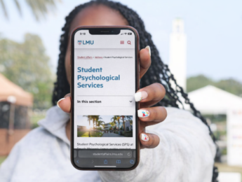 A student stands outside in front of Sacred Heart Chapel holding a phone with the SPS website on it.