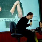 Dimitris Papaioannou speaking into a microphone on stage with Professor Katarina Zacharia smiling