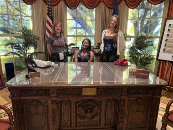 Three women from LMU Debate team sit at a large presidential desk