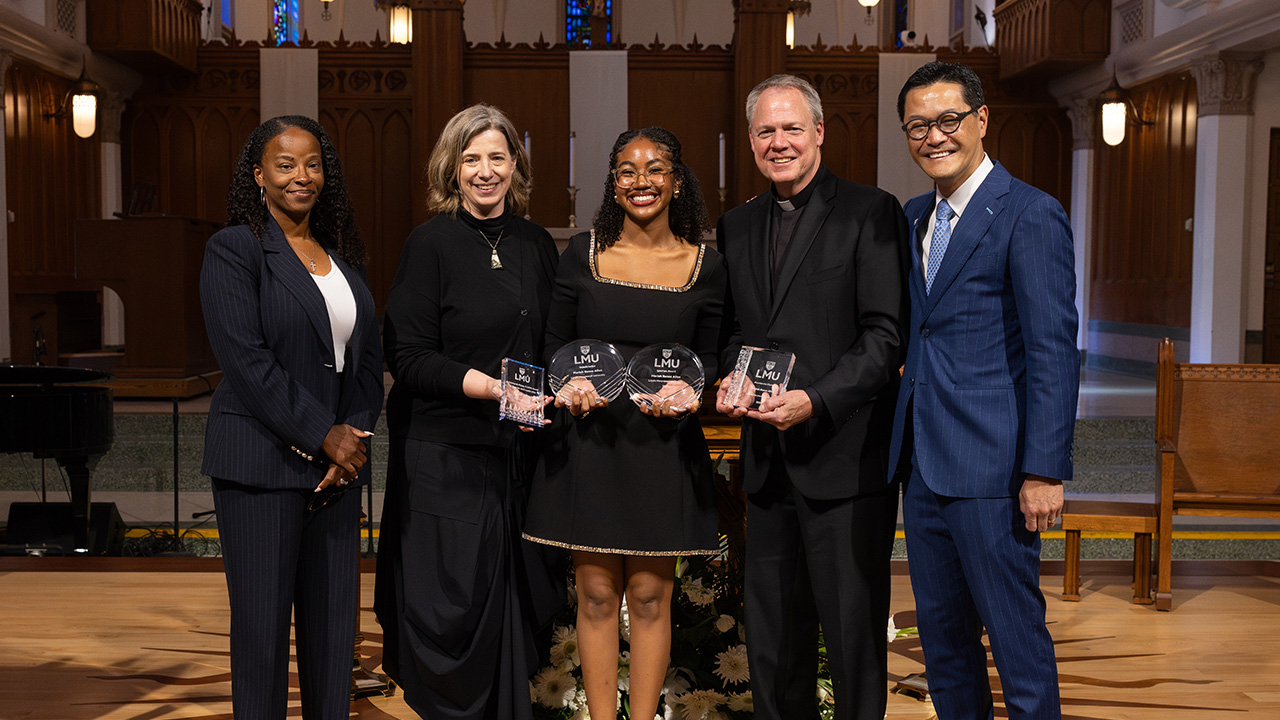 A student wearing a black dress poses with staff and administrators to receive the Class of 2026 Valedictorian and other awards at the annual student award ceremony