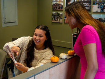 A student employee in a white sweater sits at a desk helping to give directions to another student in a pink shirt.