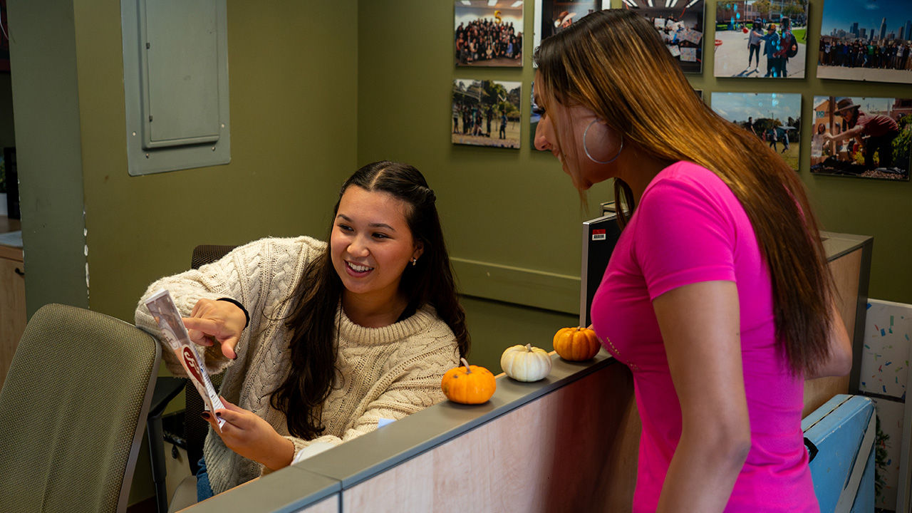 A student employee in a white sweater sits at a desk helping to give directions to another student in a pink shirt.