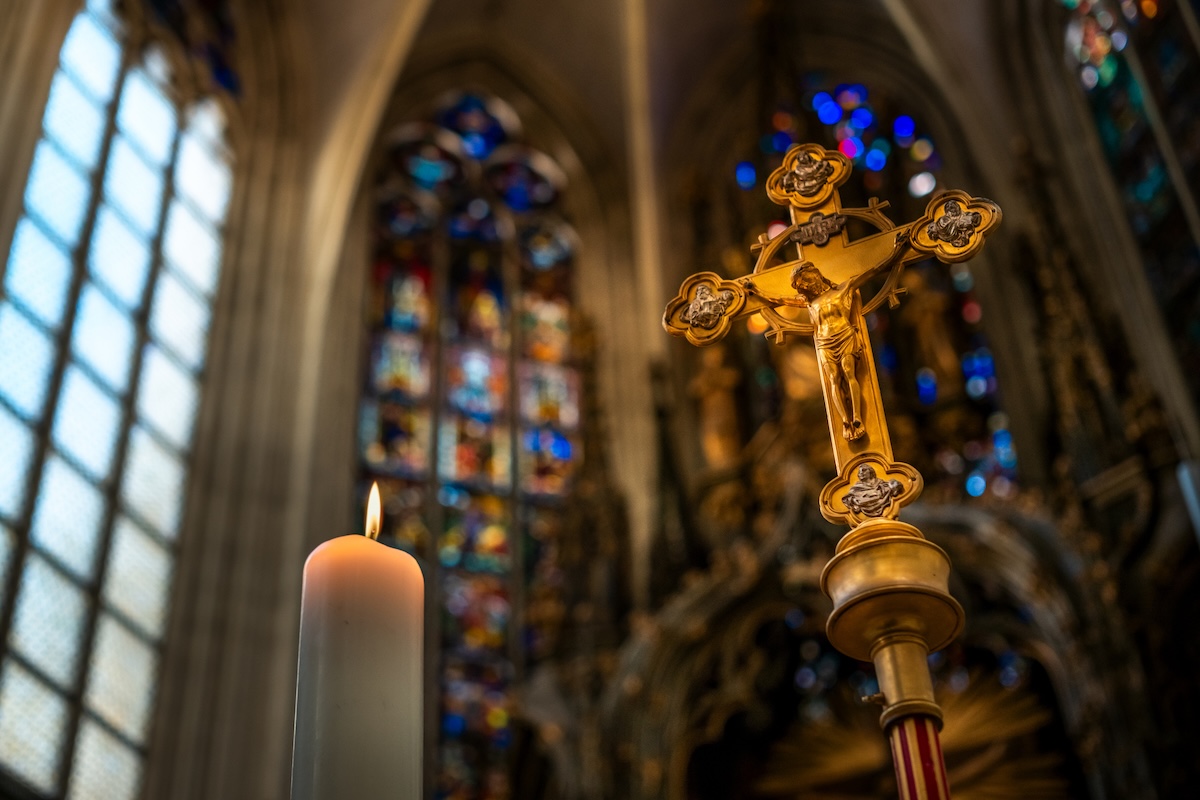 Close-up of ornate golden crucifix in a church interior, with blurred stained glass windows and vaulted ceiling in the background.