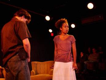 Two individuals are on a dimly lit stage performing. One is facing away, wearing a dark shirt and jeans. The other is looking off stage, wearing a light colored top and skirt. In the background, people are seated, observing the scene.