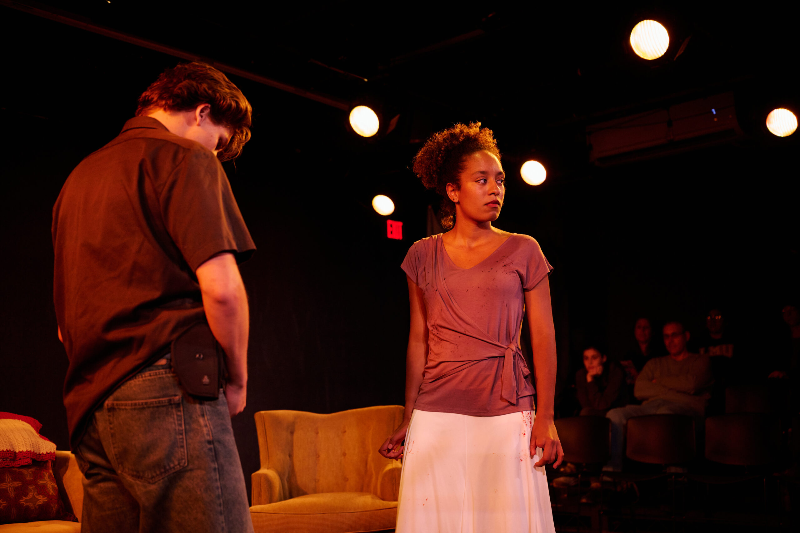 Two individuals are on a dimly lit stage performing. One is facing away, wearing a dark shirt and jeans. The other is looking off stage, wearing a light colored top and skirt. In the background, people are seated, observing the scene.