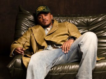 Khris is sitting on the couch, wearing a baseball hat and a tan jacket