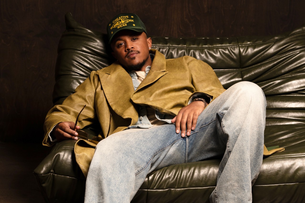 Khris is sitting on the couch, wearing a baseball hat and a tan jacket