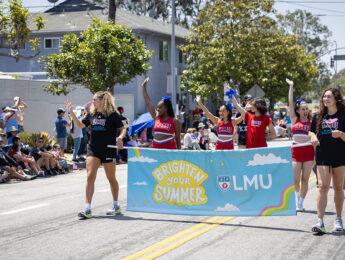 A group of LMU students walk in a Fourth of July parade carrying a banner that reads Brighten Your Summer with the LMU logo while spectators line the street in the background