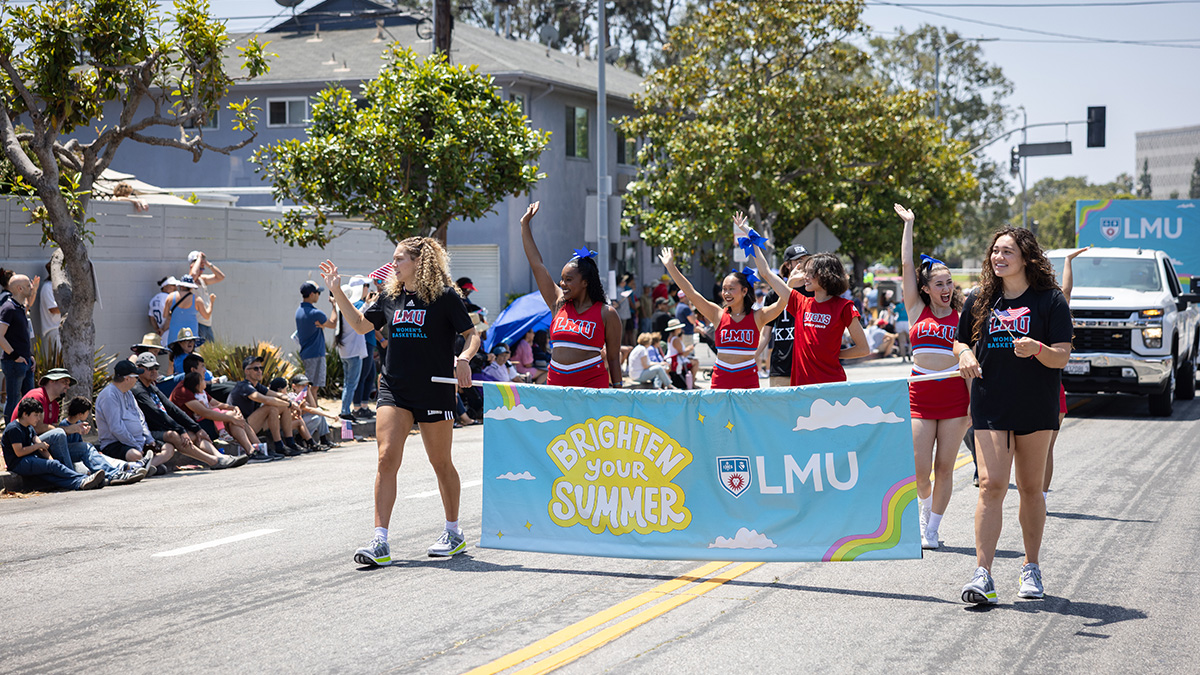 A group of LMU students walk in a Fourth of July parade carrying a banner that reads Brighten Your Summer with the LMU logo while spectators line the street in the background