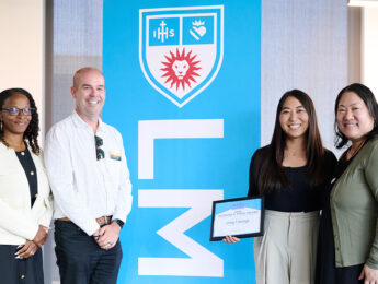 Four people stand indoors in front of an LMU banner, with one person holding a framed award certificate during a campus recognition event
