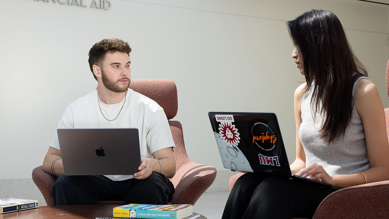 Two students sit in upholstered lounge chairs in a campus common area each working on a laptop