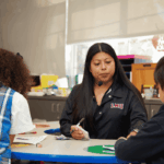Teacher in a black sweatshirt sits facing two students at a table and helps them do their schoolwork