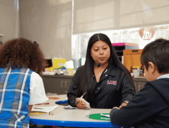 Teacher in a black sweatshirt sits facing two students at a table and helps them do their schoolwork