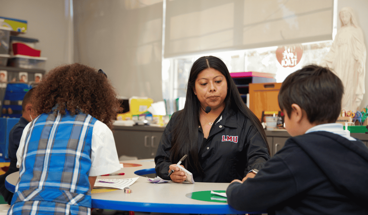 Teacher in a black sweatshirt sits facing two students at a table and helps them do their schoolwork
