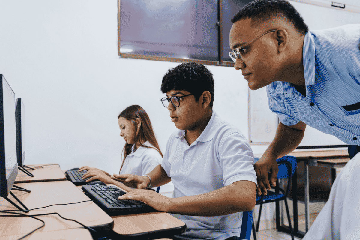 Male teacher at a Catholic school stands behind a table where two high school students are working at computers