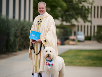 A person wearing a cream-colored ceremonial robe stands on a campus walkway holding a blue booklet and a leash attached to a large white dog
