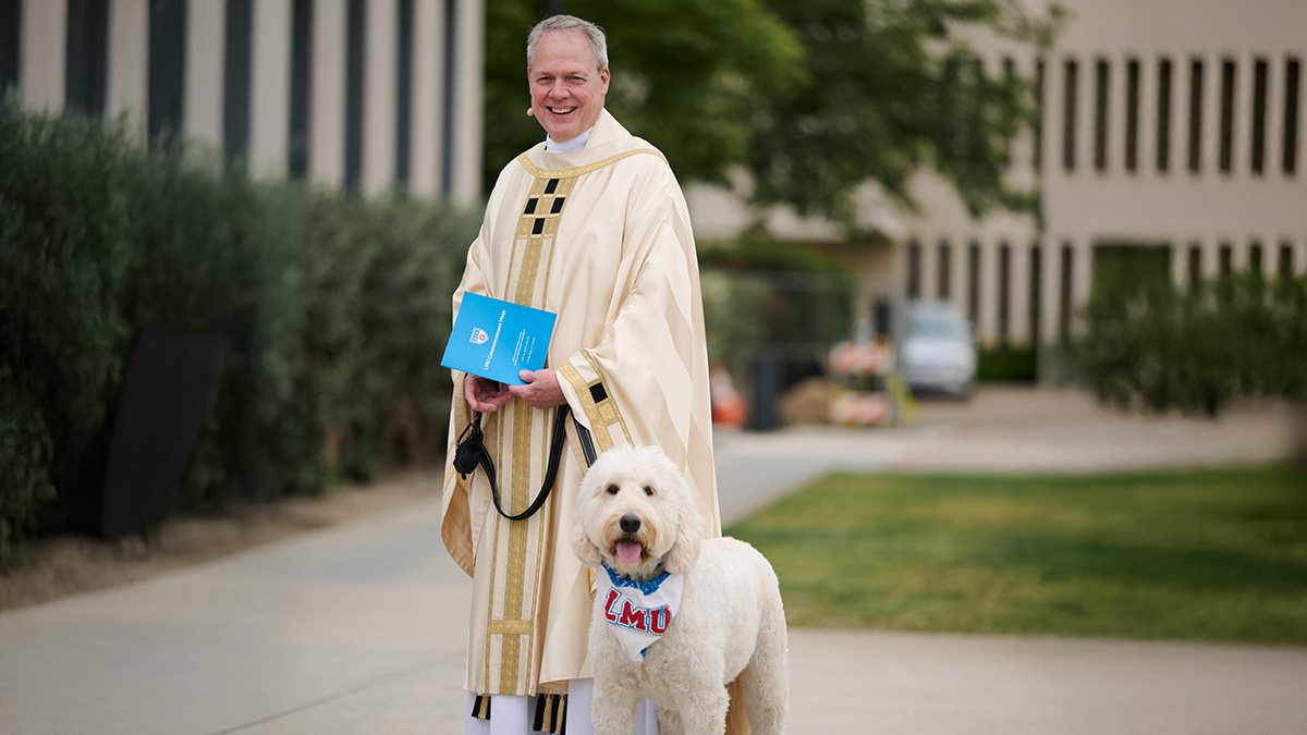 A person wearing a cream-colored ceremonial robe stands on a campus walkway holding a blue booklet and a leash attached to a large white dog