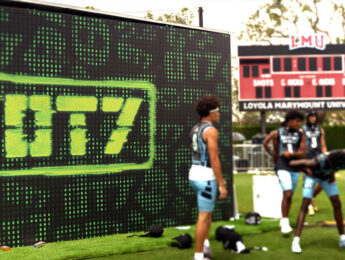 Several athletes stand on a field beside a large digital display featuring the OT7 logo