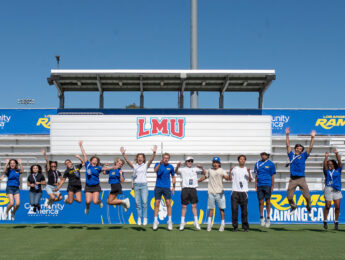 A group of approximately 15 people jump in the air on a grassy athletic field in front of stadium bleachers